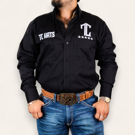 A man models the TC Hats - Black & White shirt with logo embroidery, paired with blue jeans, a brown tooled leather belt, gold watch, and jewelry against a plain white background.