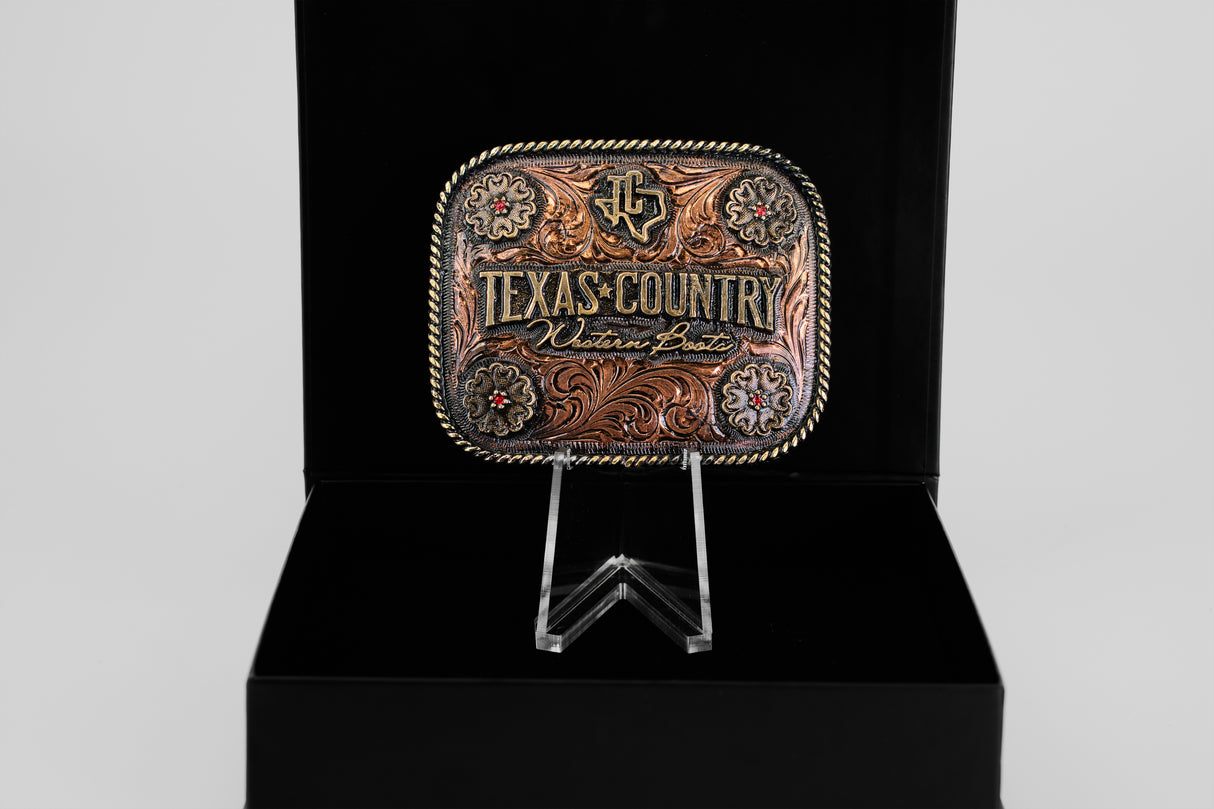 The Texas Country Buckle - Mod 2 features gold and brown details with “Texas Country” and “Western Boots” engraved, displayed upright on a clear stand against a black background.