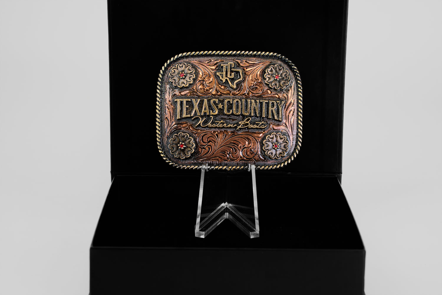 The Texas Country Buckle - Mod 2 features gold and brown details with “Texas Country” and “Western Boots” engraved, displayed upright on a clear stand against a black background.