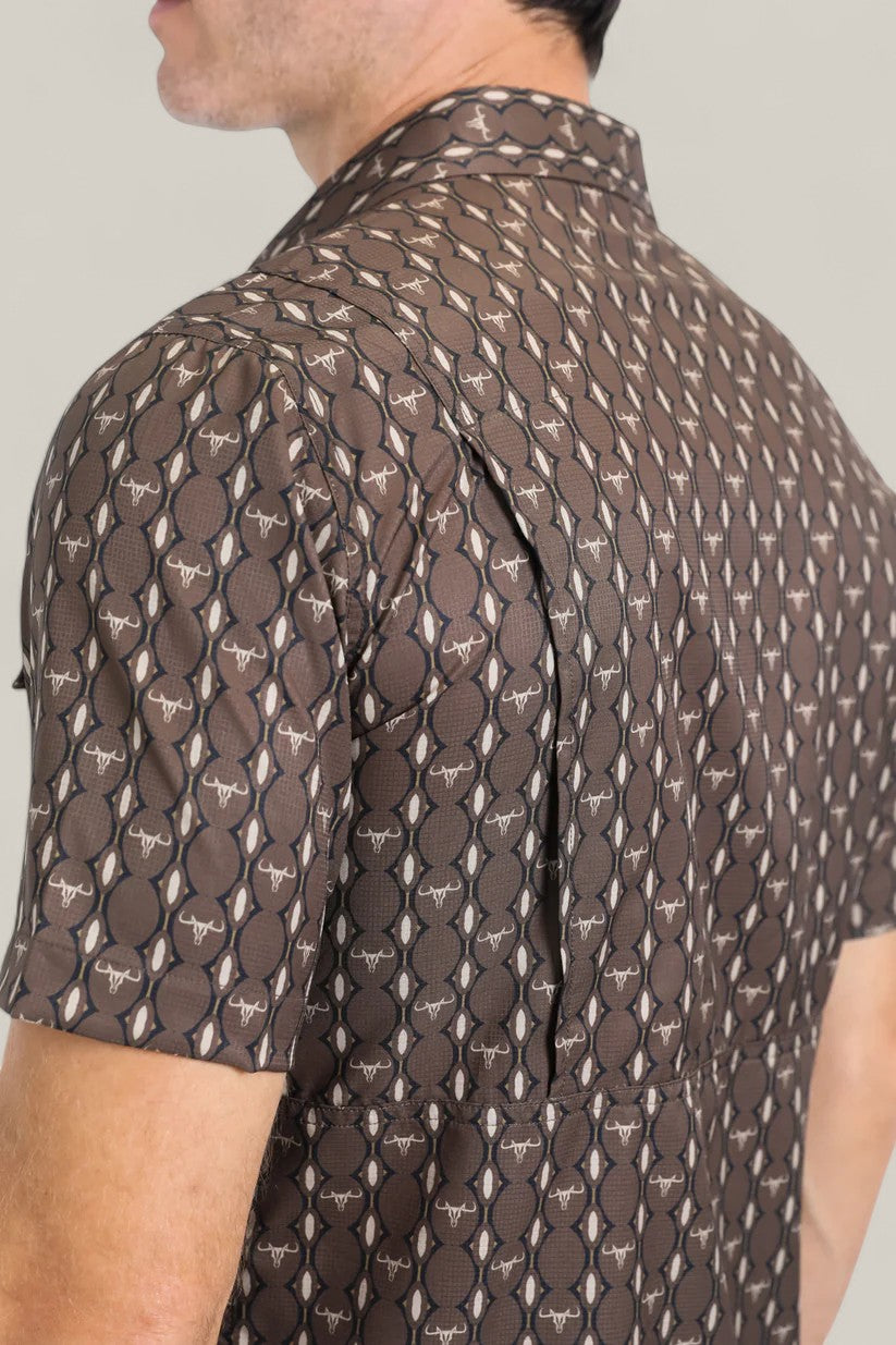 Close-up of a man wearing the Mens Performance Western Short Sleeve Aztec Print Brown Shirt (PGW10643), featuring brown and beige Aztec patterns with white bull head motifs. Only his upper back and part of his face are visible.