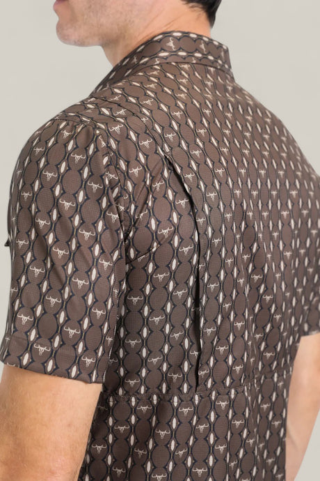 Close-up of a man wearing the Mens Performance Western Short Sleeve Aztec Print Brown Shirt (PGW10643), featuring brown and beige Aztec patterns with white bull head motifs. Only his upper back and part of his face are visible.