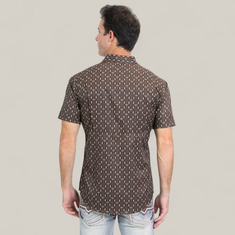 A man stands with his back to the camera, wearing a Mens Performance Western Short Sleeve Aztec Print Brown Shirt - PGW10643 and light blue jeans with decorative pocket stitching, set against a plain light background.
