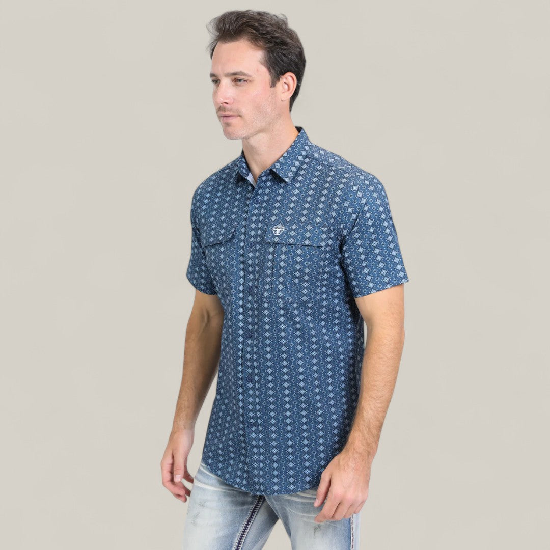 A man wearing the Mens Performance Western Short Sleeve Aztec Print Navy Shirt (PGS10659) with light blue jeans stands against a plain, light gray background, facing slightly right with a neutral expression.