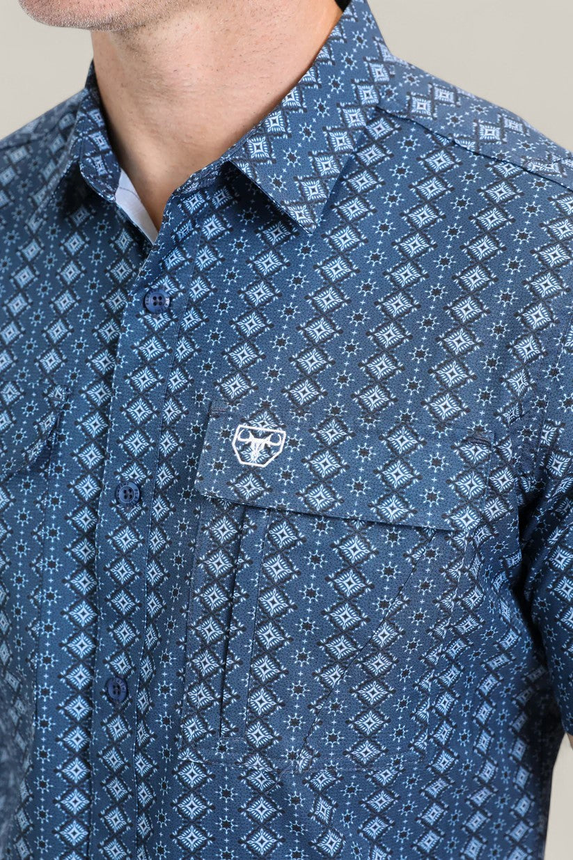 A man wears the Mens Performance Western Short Sleeve Aztec Print Navy Shirt (PGS10659), showcasing its geometric Aztec design and chest pocket with a white embroidered logo. Only his lower face and upper torso are visible.