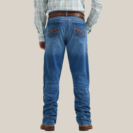 A person wearing Wrangler 20x Mens Medium Wash Backwater Vintage Bootcut Stretch Denim Jeans with a brown tooled leather belt, plaid shirt, and brown cowboy boots. The image highlights the lower half of the body against a plain background.