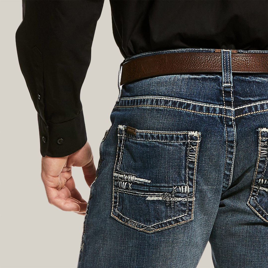 A person is wearing M5 Slim Stretch Adkins Stackable Straight Leg Jean (lennox - 10030275) with frayed back pockets, a brown leather belt, and a black shirt with buttoned cuffs. Their left hand rests by their side, highlighting the jeans durable denim and stylish worn look.