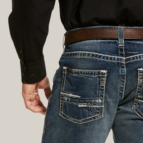 A person is wearing M5 Slim Stretch Adkins Stackable Straight Leg Jean (lennox - 10030275) with frayed back pockets, a brown leather belt, and a black shirt with buttoned cuffs. Their left hand rests by their side, highlighting the jeans durable denim and stylish worn look.
