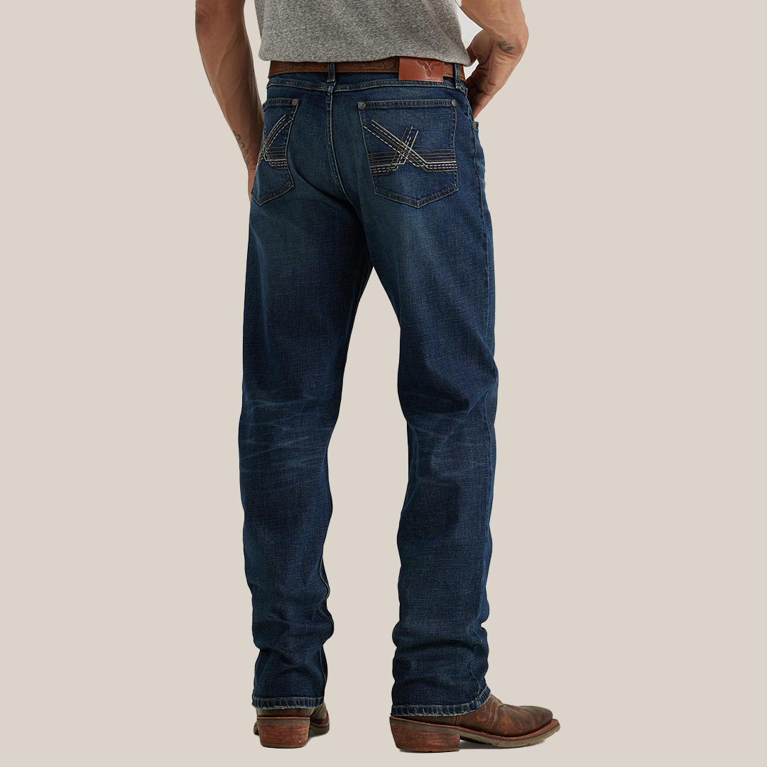 A person in a gray shirt and Wrangler 20X Sunnybrook Dark Wash Extreme Relaxed Straight Stretch Denim Jeans with decorative back pocket stitching stands on a plain white background. They complement the look with brown leather boots and a matching belt, highlighting the jeans relaxed fit.