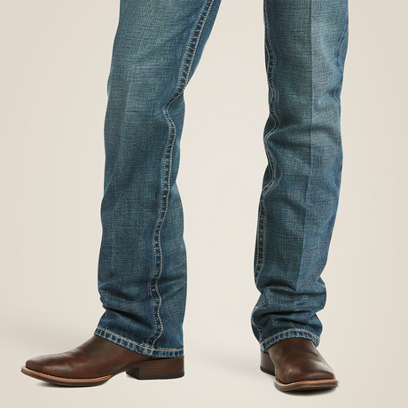 A person is wearing a dark gray t-shirt, Ariat M5 Slim Straight Leg Jeans in blue denim (Gulch - 10014010), and brown leather boots. They have a brown belt with a silver buckle and are standing against a white background with one hand in their pocket.
