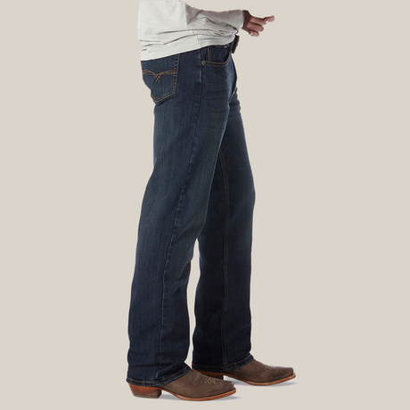 A person wearing Wrangler 20X No. 33 Extreme Appleby Dark Wash Relaxed Straight Leg Jeans and brown cowboy boots, hands on their waist. The jeans relaxed fit blends effortlessly with the plain light gray background.