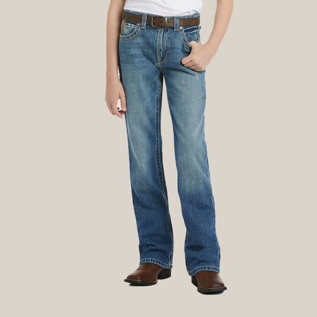A young boy stands with legs slightly apart, wearing Ariats medium wash boot cut jeans with a faded look and brown leather cowboy boots featuring a pointed toe and block heel, set against a white background.