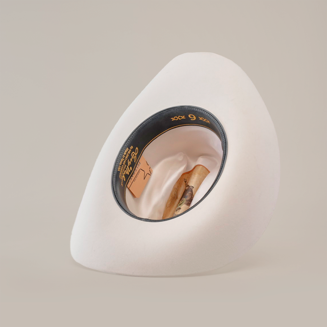The 6X Real White cowboy hat, expertly crafted by artisans from fur felt, sits at an angle, revealing its black inner band. Nestled inside this luxury Western headwear is a rolled cigar. The studio backdrop is plain white.
