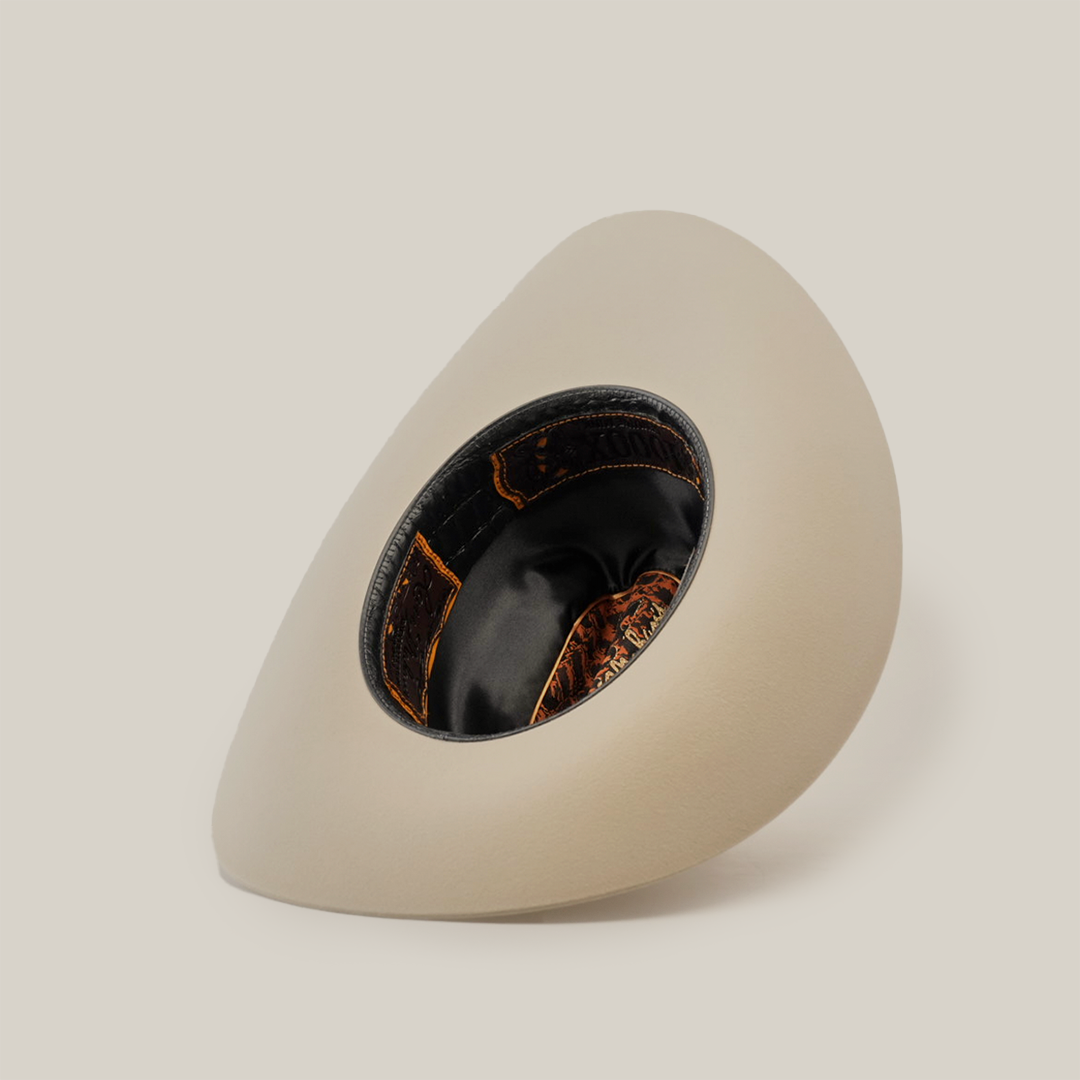 A tilted shot of a white wide-brimmed western hat highlights its interior, featuring a shiny black band and patterned brown and orange lining, reminiscent of the classy 1000X Imperial Belly collection. The softly lit background is plain.