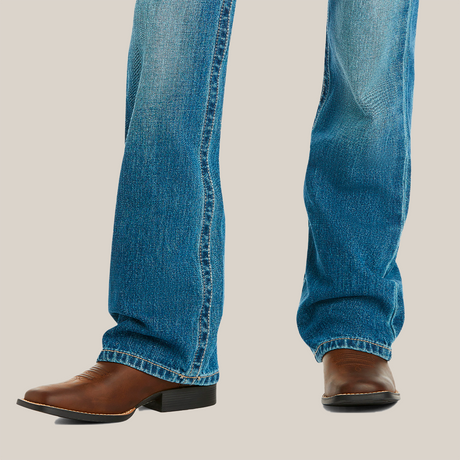 A young boy stands with legs slightly apart, wearing Ariats medium wash boot cut jeans with a faded look and brown leather cowboy boots featuring a pointed toe and block heel, set against a white background.