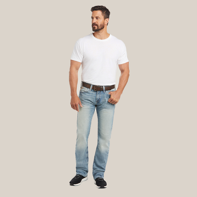 Against a plain background, a man with a beard and short hair wears a white T-shirt and light blue M7 Rocker Stretch Stirling Stackable Straight Leg Jean by Ariat in Shasta. He complements his outfit with a brown belt and black sneakers, looking to his left with his hands by his sides.