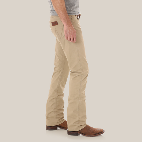 Wearing Mens Wrangler Retro Slim Fit Straight Leg Pants 1088MWZFN with a brown leather patch on the pocket, a gray shirt, and brown cowboy boots, they stand sideways against a white background, capturing the classic Wrangler Retro style.