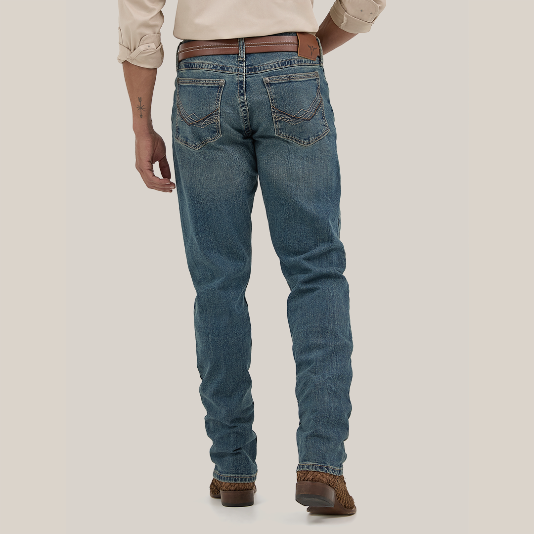 A person in Wrangler Mens 20X Extreme Relaxed Fit Jeans - 1044MWXMA, blue denim with a brown belt and shoes, stands against a white background. They sport an arm tattoo and light-colored rolled sleeves, epitomizing the relaxed style.