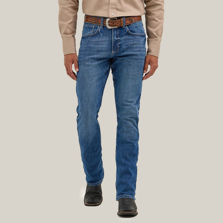 A person in a beige long-sleeve shirt, blue jeans, and a brown belt stands before a plain background. They wear Mens Wrangler 20X No. 42 Vintage Bootcut Jeans with the iconic patch, hands at their sides, while black boots complete the outfit.