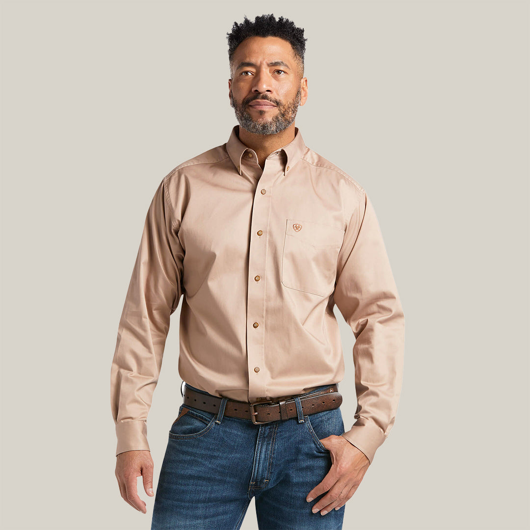 A man with short curly hair and a beard wears the Solid Twill Classic Fit Shirt Kaki - 10000505, featuring elegant embroidery. Paired with a brown belt and jeans, he stands against a plain white backdrop, exuding timeless style.
