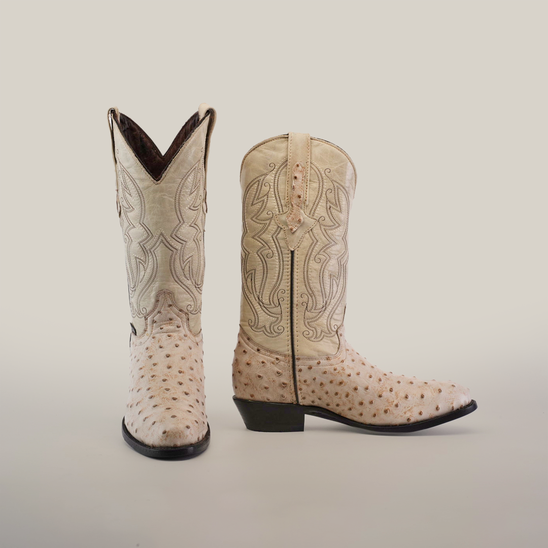 The Karoo Ostrich Print Bone boots, in a light color, feature intricate stitching and an ostrich print texture. One boot stands upright while the other lies on its side to showcase its distinctive heel and side design.