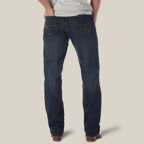 A person poses against a white backdrop wearing the Wrangler 20X No. 33 Extreme Appleby Dark Wash Relaxed Straight Leg Jeans, featuring orange stitching and styled with brown shoes, embodying a laid-back essence with hands in back pockets and a light gray shirt.