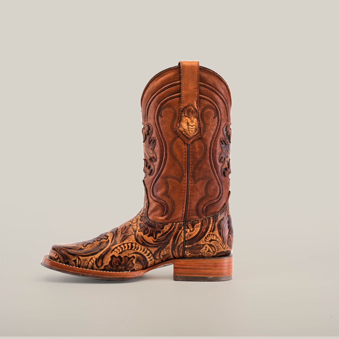 The Handtooled Leave Orix cowboy boot, made of premium brown cowhide leather, features intricate floral and swirl embossing on the shaft and foot. With a low wooden heel, rodeo toe, and slip-resistant sole, it combines style with functionality against a plain white backdrop.