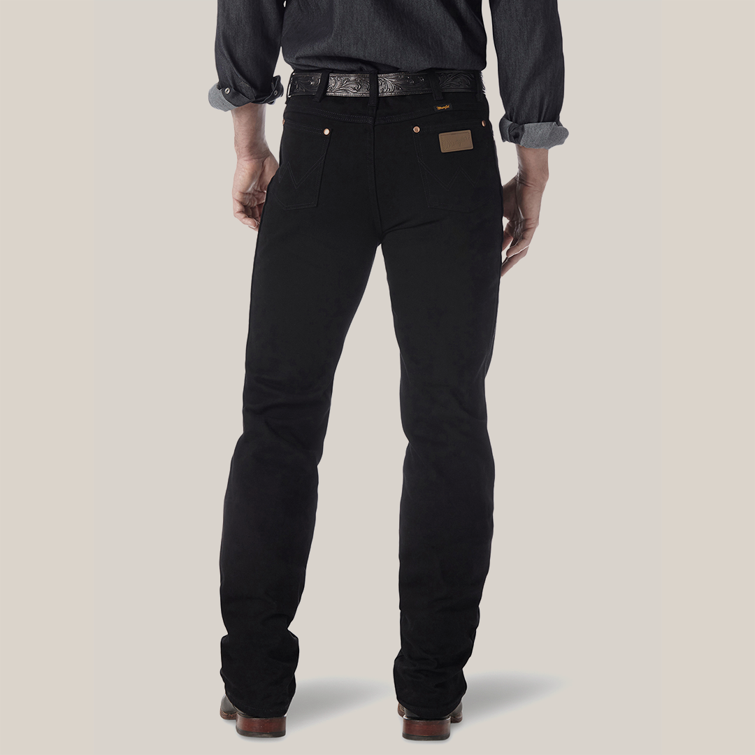 A person faces away from the camera, wearing a dark long-sleeve shirt with rolled-up sleeves, black Wrangler Cowboy Cut Slim Fit Jeans - 0936WBK made from premium denim, and brown leather shoes. A belt is partially visible above the waistband of their jeans.
