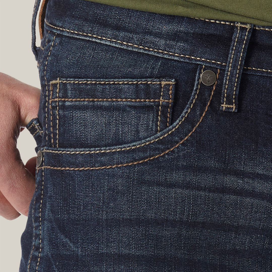 A person wearing Mens Wrangler 20X No. 42 Vintage Bootcut Jean - 42MWXRD in premium dark blue denim, accented with orange stitching. With a hand adorned by a silver ring tucked into the pocket and paired with a green shirt, the stylish slim fit jeans exude comfort.