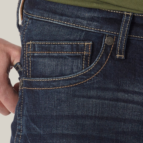A person wearing Mens Wrangler 20X No. 42 Vintage Bootcut Jean - 42MWXRD in premium dark blue denim, accented with orange stitching. With a hand adorned by a silver ring tucked into the pocket and paired with a green shirt, the stylish slim fit jeans exude comfort.