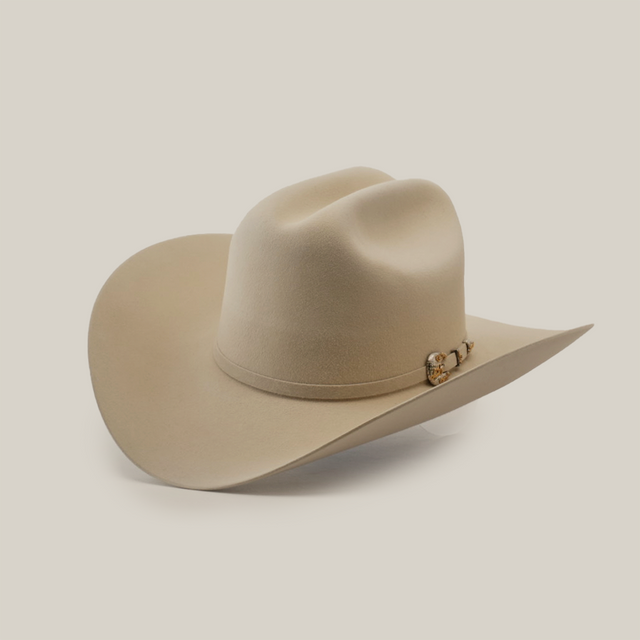 The 1000X Imperial Belly hat is a beige cowboy style with a wide brim, adorned with a decorative band featuring a small metallic emblem, set against a plain white background.