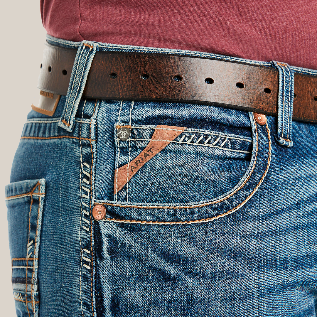 A close-up of someone wearing M5 Slim Stretch Stillwell Stackable Straight Leg Jeans - Fargo shows premium cotton blue denim with detailed stitching and a pocket logo. A brown leather belt with metal holes loops through the belt loops, effortlessly complementing the maroon shirt.