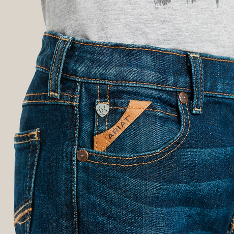 A person is wearing Ariat Boys B4 Legacy Dark Wash Relaxed Fit Boot Cut Jeans, featuring yellow stitching, a small front pocket with a brown leather Ariat label, and a silver button on the waistband. A gray shirt peeks out above the jeans.