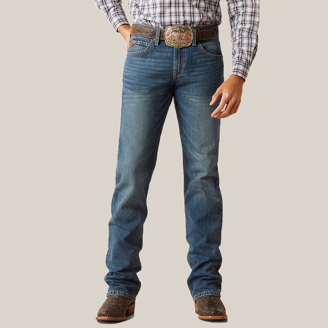 Dressed in the M7 Slim Ezra Straight Jean - Brighton, a person stands with hands at sides. The jeans have a slightly faded look, ideal for a modern cowboy, paired with brown cowboy boots and a large decorative belt buckle.