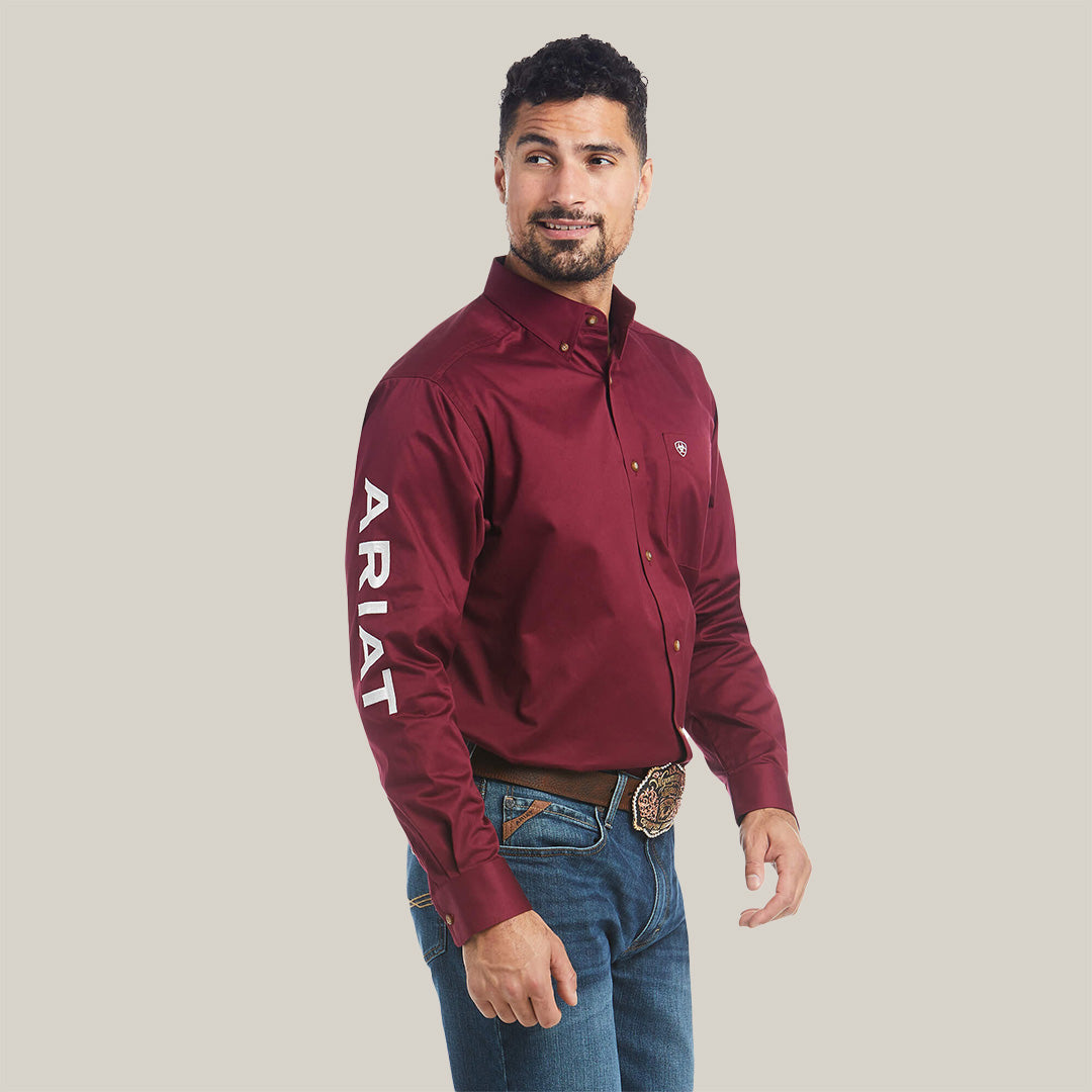 A man in a Team Logo Twill Classic Fit Shirt Burgundy - 10027995, featuring ARIAT on the right sleeve, blue jeans, and a decorative belt buckle, stands against a white background. His short dark hair and beard enhance his relaxed expression as he gazes to the side.