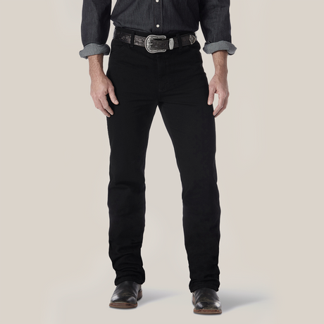 Someone wearing sleek Wrangler Cowboy Cut Slim Fit Jeans, a dark button-up shirt with rolled sleeves, paired with a decorative belt featuring a silver buckle, and stylish dark leather shoes, stands against a plain light-colored background that highlights the ensembles modern appeal.