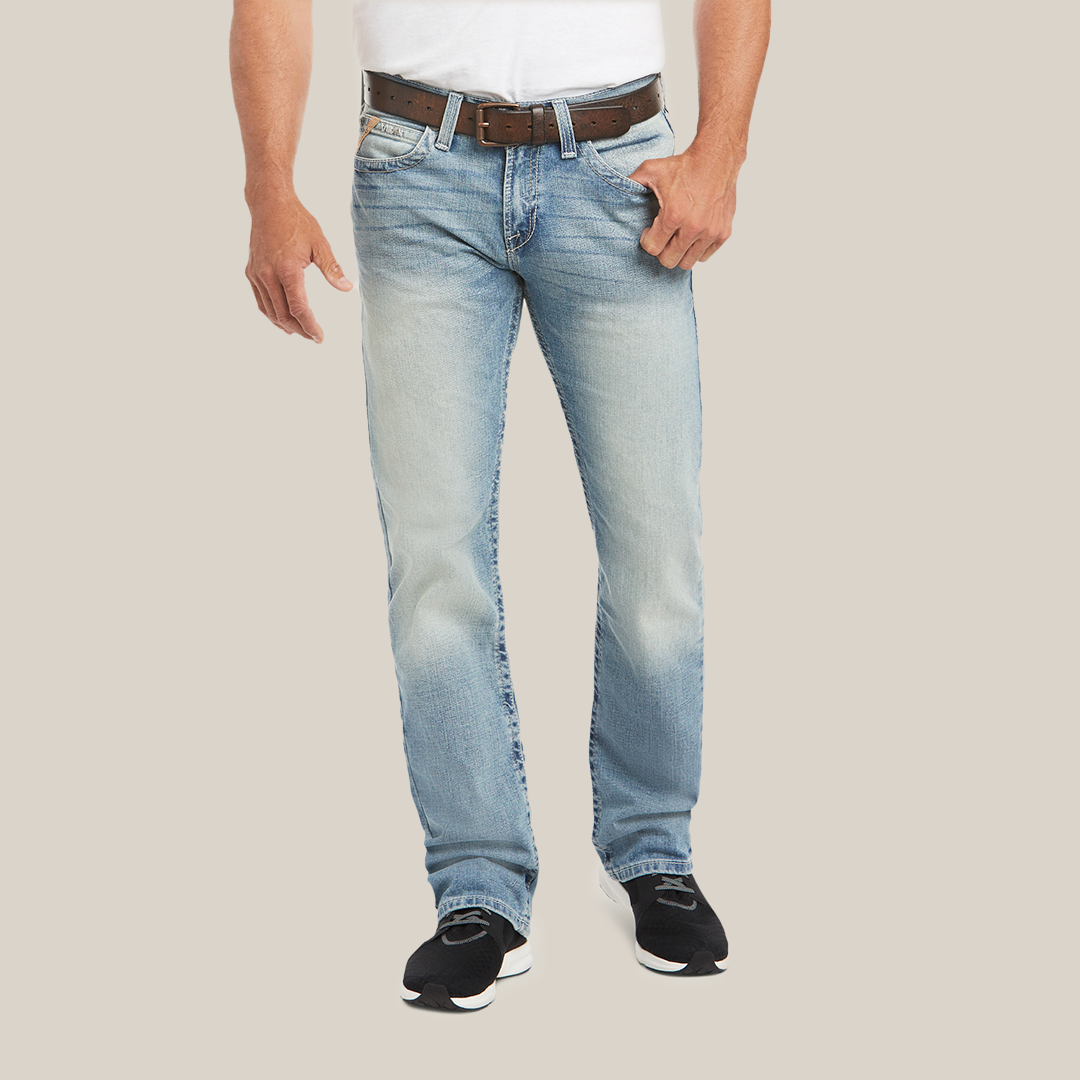 Someone in a white shirt, light blue M7 Rocker Stretch Stirling Stackable Straight Leg Jean - Shasta (10031997), and black shoes stands against a plain backdrop. The slightly faded jeans with stretch fabric are complemented by the brown belt held in one hand. The image is cropped at the shoulders.