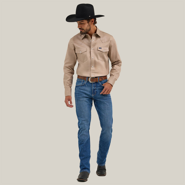 A man in a cowboy hat stands against a plain white background, wearing a beige long-sleeved shirt and Mens Wrangler 20X No. 42 Vintage Bootcut Jeans crafted from premium denim. Dark boots complete his rugged look as he gazes downward, embodying timeless Western style.