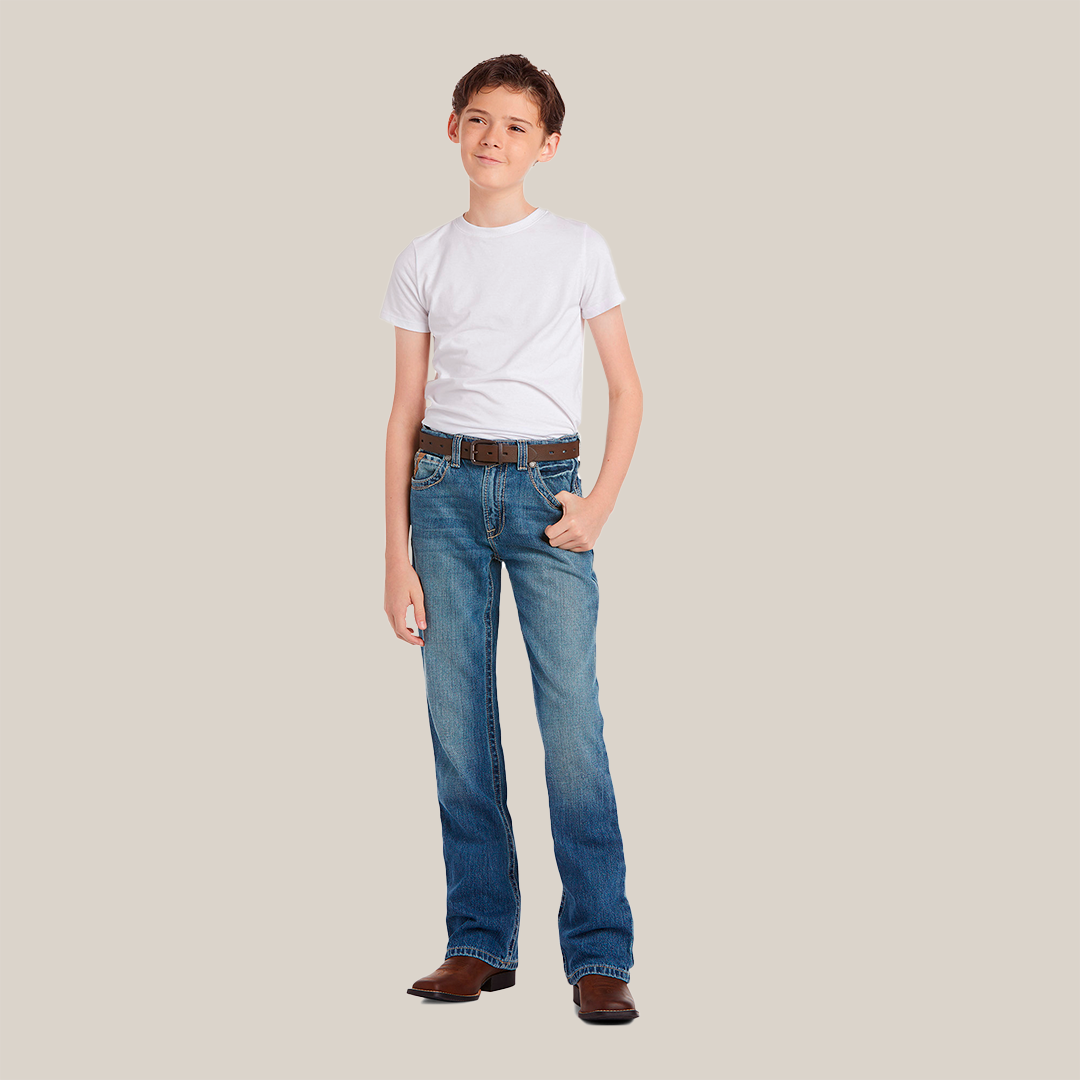 A person leans casually against a plain beige backdrop, wearing a white shirt, Ariat Boys B4 Boundary Dakota Medium Wash Relaxed Fit Boot Cut Jeans - 10018345, brown cowboy boots, and a matching brown belt with one hand in their pocket.