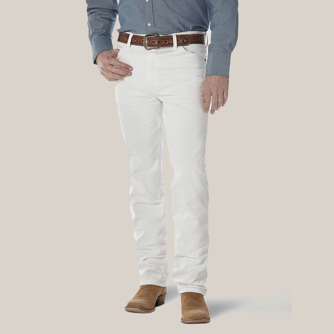 A person wearing a blue button-up shirt, Wrangler Cowboy Cut Slim Fit Jeans (0936WHI), and a brown leather belt is grounded in brown suede shoes. The iconic Wrangler patch enhances the look against the plain white background.