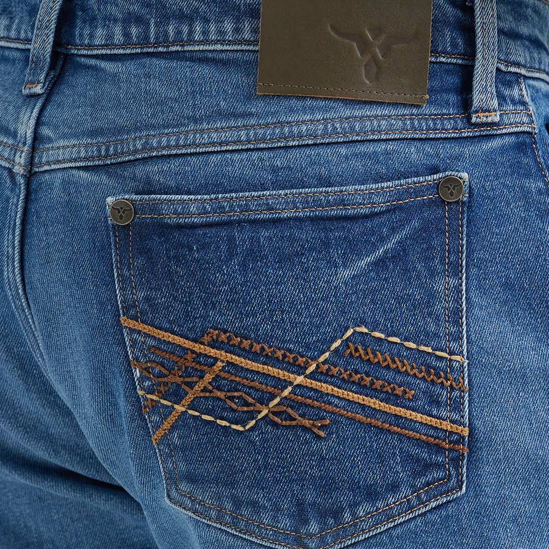 A close-up shows the back pocket of Wrangler 20x Mens Medium Wash Backwater Vintage Stretch Jeans, featuring an orange and brown embroidered geometric design. A logo leather patch is on the waistband. The jeans pair classically with a tucked plaid shirt for a timeless style.