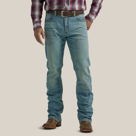 A person in Wrangler 20X Mens Light Wash Shade Bootcut Stretch Jeans and a plaid shirt stands facing away. The premium denim jeans boast decorative stitching on the back pockets, complemented by a brown belt and shoes, set against a plain, neutral backdrop.