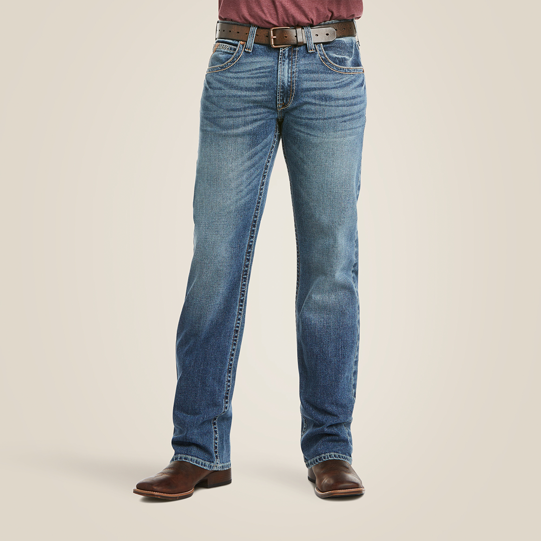 Wearing M5 Slim Stretch Stillwell Stackable Straight Leg Jean - Fargo, paired with brown leather boots, a brown belt, and a maroon shirt, a person stands against a plain background. The faded jeans offer a worn-in look for both comfort and style.