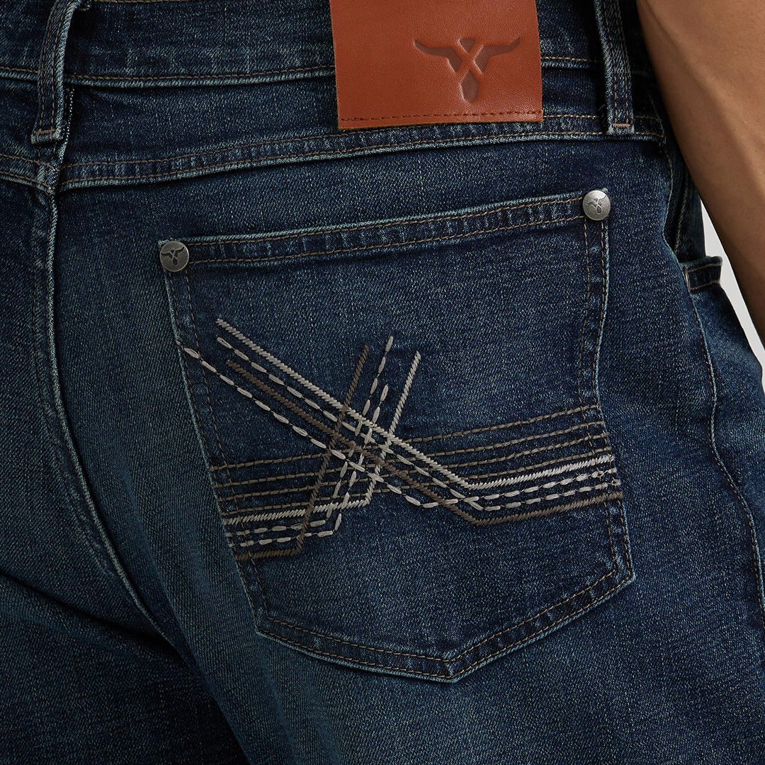 A person wears Wrangler 20X Sunnybrook Dark Wash Extreme Relaxed Straight Stretch Denim Jeans, paired with a gray shirt. The premium jeans feature decorative stitching on the back pocket and a brown leather patch with a small logo on the waistband.