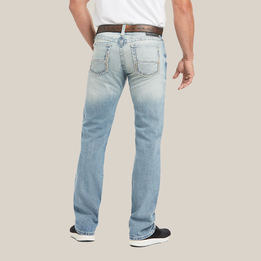 Rear view of a person in M7 Rocker Stretch Stirling Stackable Straight Leg Jeans - Shasta, displaying light blue faded stretch fabric. They wear a brown belt and white shirt, stand with one leg bent in black and white sneakers, emphasizing the relaxed straight-leg fit.