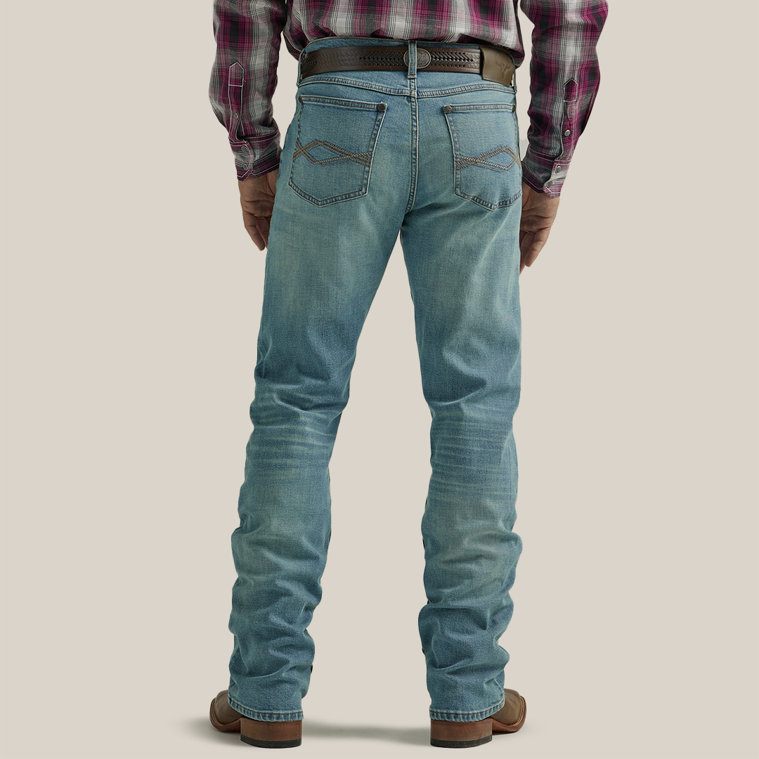 A close-up shows a person in Wrangler 20X Mens Light Wash Shade Bootcut Stretch Jeans (112335407) paired with a brown woven leather belt featuring a decorative round buckle, and a neatly tucked plaid shirt.