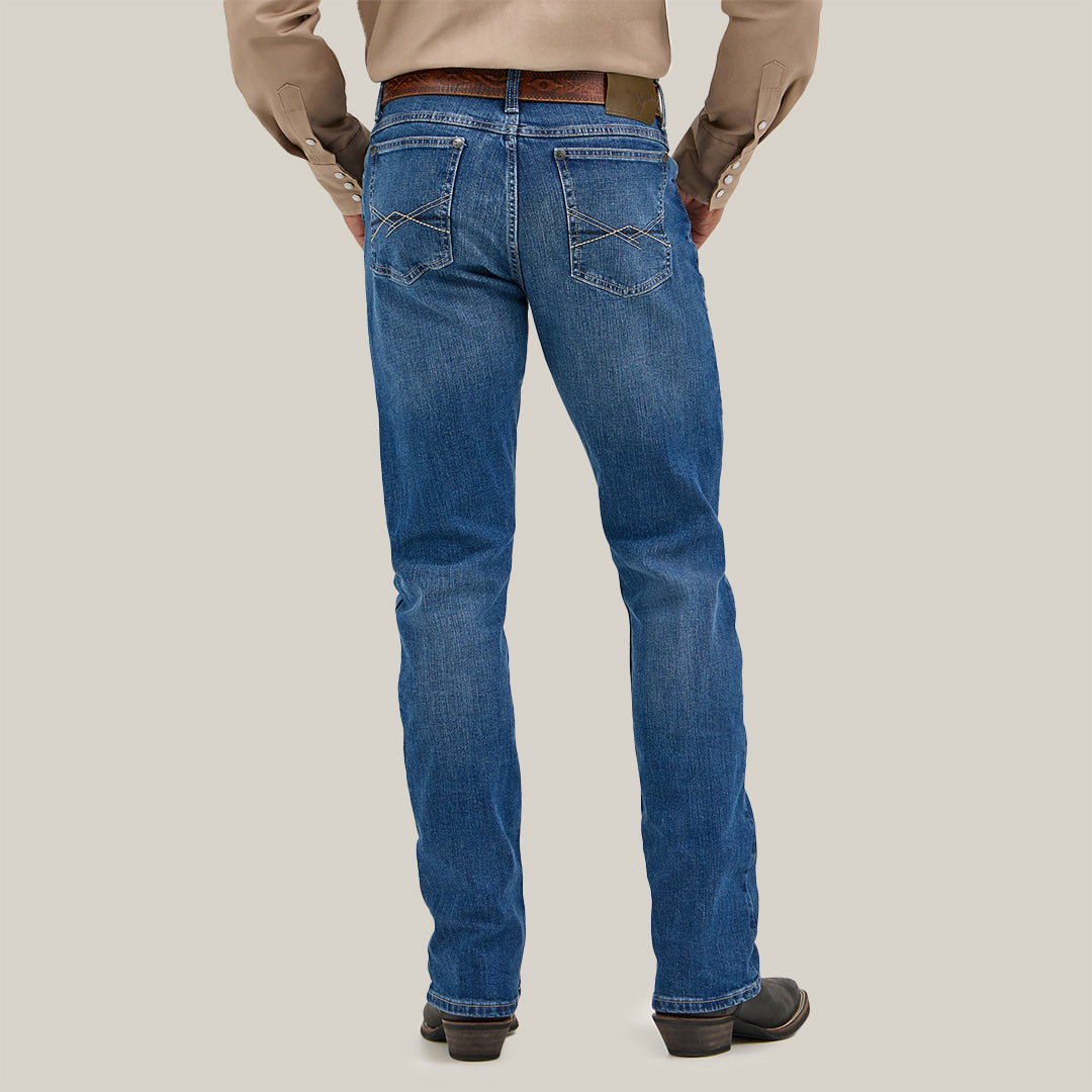 A person in bootcut jeans wears a beige long-sleeve shirt, seen from the back. The Wrangler 20X No. 42 features premium denim with a classic design and a Wrangler patch on the pocket. Black boots and a brown belt complete the look against a plain, light background.