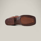 The image features the sole of a Grisly Tan - Square Toe boot, made of premium leather with stitching and small perforations. This Western-style shoe has a textured grip and rectangular heel, set against a plain light gray background.