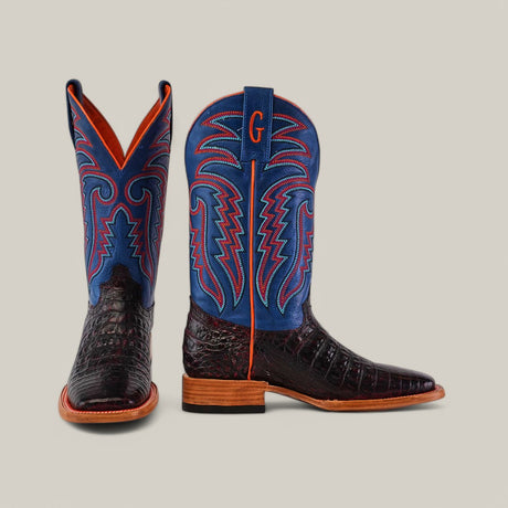 The Exotic Caiman Belly - Black Cherry boots feature dark brown, textured leather on the foot, vibrant blue shafts with intricate red and white stitching, a wooden heel, and a bold G on the striking blue shaft.