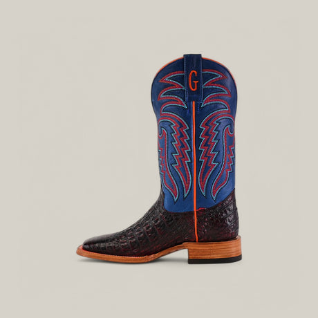 The Exotic Caiman Belly - Black Cherry cowboy boot features a dark brown crocodile-patterned lower, a blue upper with red and blue stitching, a wooden sole, and a handcrafted red G on the blue section near the top, all set against a neutral background.