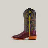 The Exotic Caiman Belly Tail boot, crafted from maroon alligator leather, showcases a blue shaft with yellow and maroon stitching and a yellow logo. It features a wooden heel and square toe design, set against a beige background.